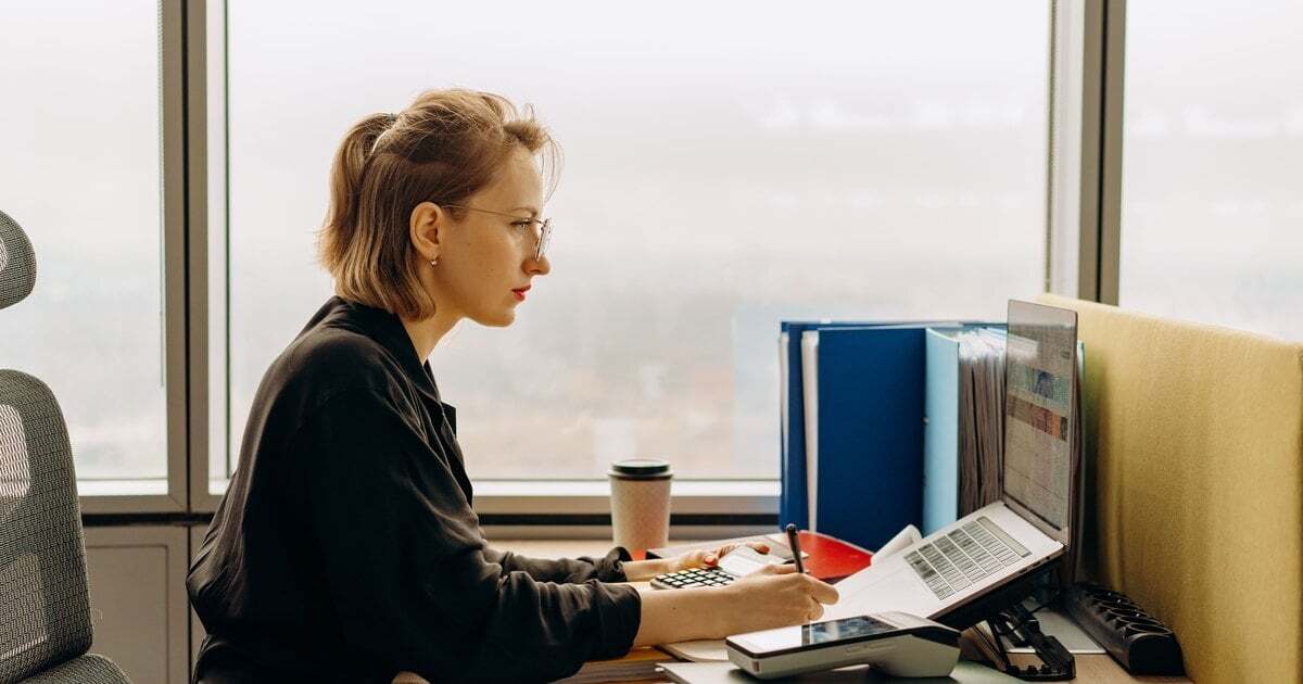 A professional at a desk by a window, working through figures with a calculator and laptop