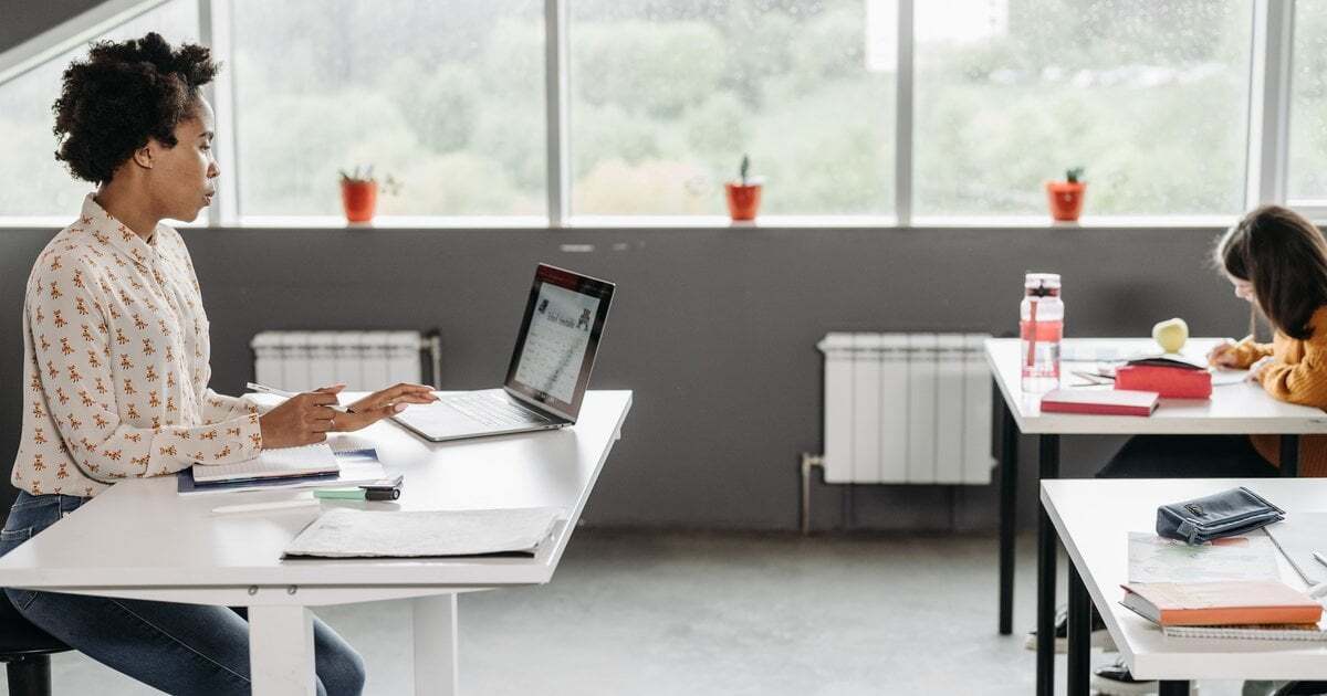A teacher working on a laptop at the front of a classroom while a student writes at a desk