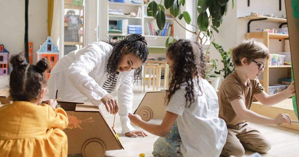 An educator crouches to help young children paint cardboard cars on a playroom floor