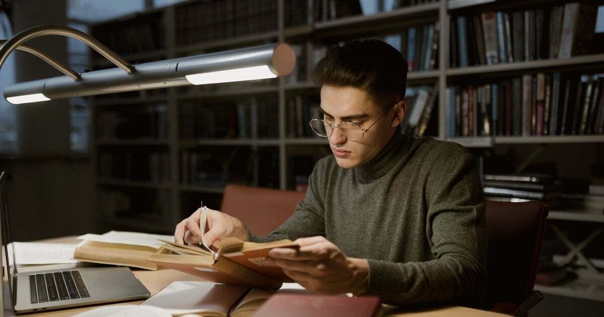 A student reading in a library at night, open books and a laptop on the desk