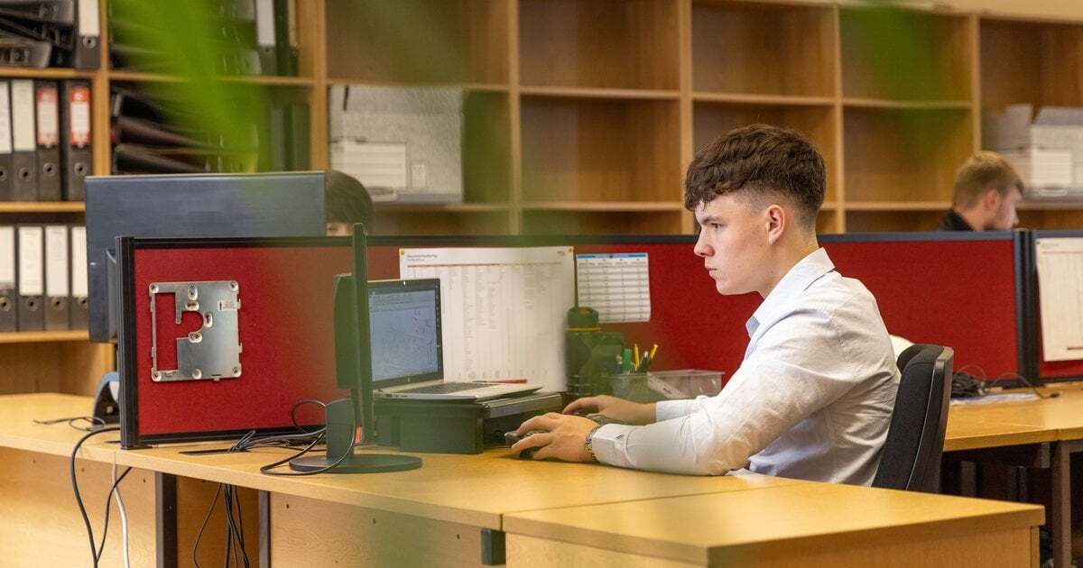 A young professional working at a computer in an office, shelves of files behind him